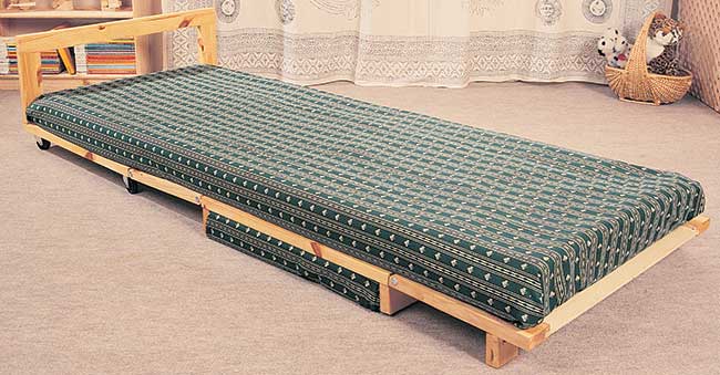 Il mobile in pratica si avvolge e si svolge come un riccio: per portarlo dalla funzione poggiapiedi (1) a quella letto (3) basta sollevare il cuscino che (2) si tira dietro la coda del letto che pendeva appoggiata contro la sua spalliera. Svolto, il letto poggia sulle quattro ruote della parte del capezzale, sulle sponde che ospitano il cuscino e sui piedini incernierati all’elemento finale chiaramente visibili, ancora ripiegati, nella seconda foto. Le aperture frontali possono facilmente essere coperte da teli cuciti al cuscino, appesantiti con piombini.